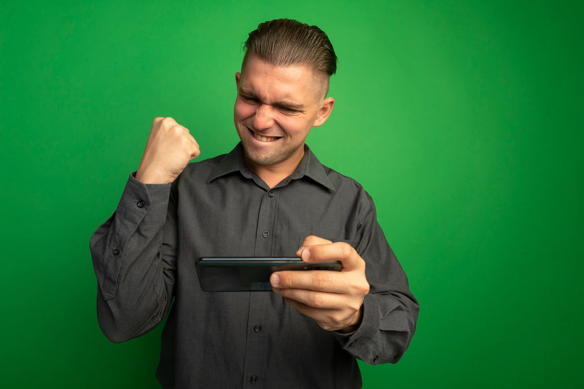 Young handsome man grey shirt using gadget clenching fist happy excited rejoicing his success standing green